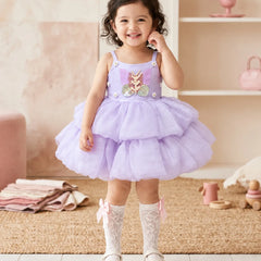 A toddler girl stands in a softly styled nursery wearing a lavender sleeveless tutu dress with a floral applique bodice, white lace socks, and white shoes.