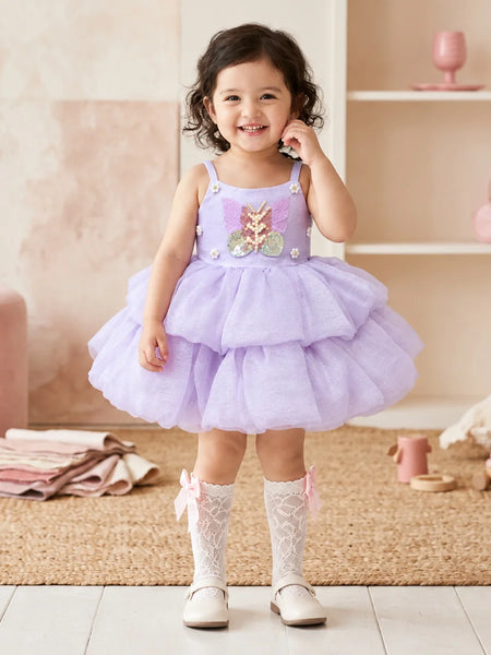 A toddler girl stands in a softly styled nursery wearing a lavender sleeveless tutu dress with a floral applique bodice, white lace socks, and white shoes.