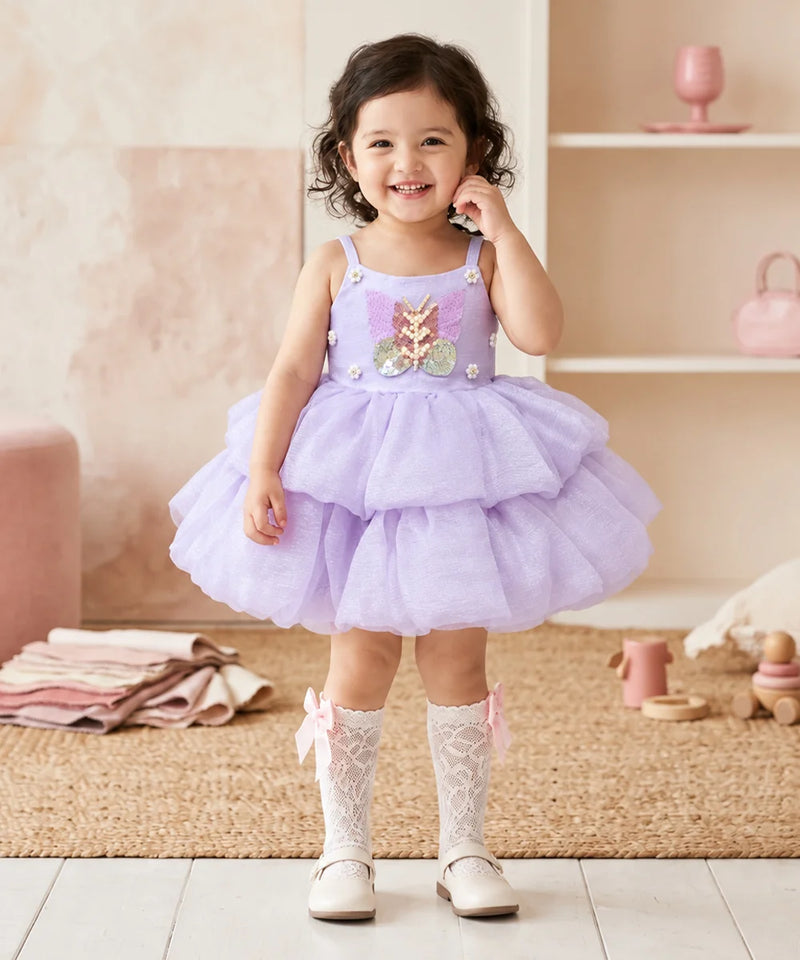 A toddler girl stands in a softly styled nursery wearing a lavender sleeveless tutu dress with a floral applique bodice, white lace socks, and white shoes.