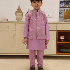 A child wearing a purple kurta pyjama set with an embellished Koti waistcoat standing against an indoor background.