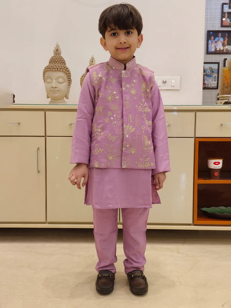 A child wearing a purple kurta pyjama set with an embellished Koti waistcoat standing against an indoor background.