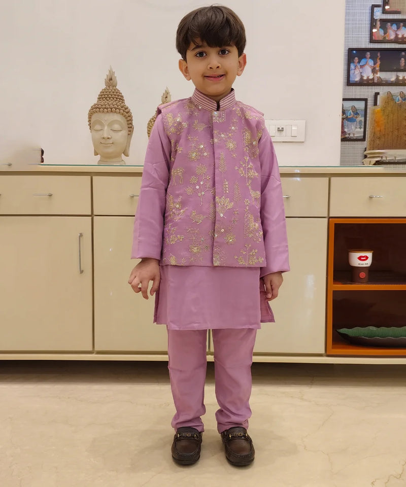 A child wearing a purple kurta pyjama set with an embellished Koti waistcoat standing against an indoor background.