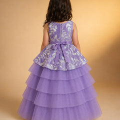 A young girl viewed from behind wearing a sleeveless lavender dress with a silver floral patterned peplum waist and a tiered tulle skirt, set against a warm beige studio background.