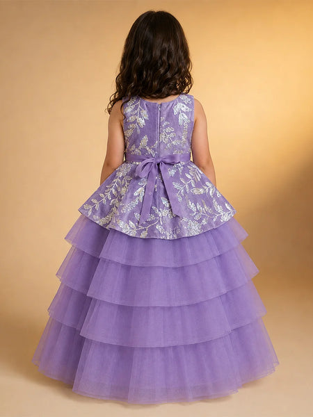 A young girl viewed from behind wearing a sleeveless lavender dress with a silver floral patterned peplum waist and a tiered tulle skirt, set against a warm beige studio background.
