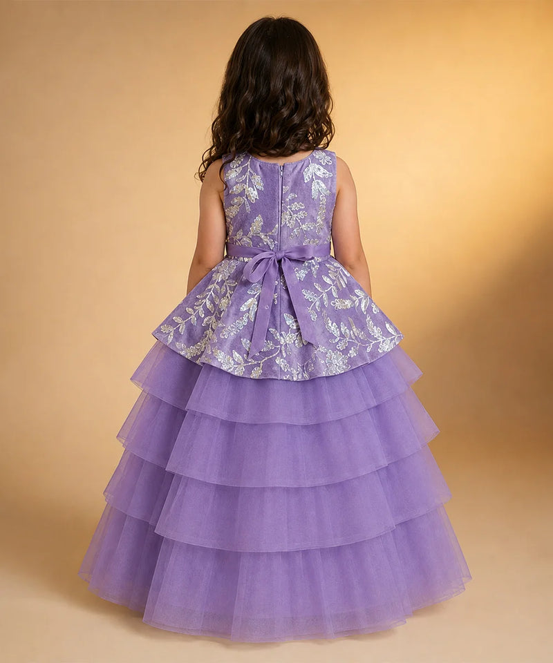 A young girl viewed from behind wearing a sleeveless lavender dress with a silver floral patterned peplum waist and a tiered tulle skirt, set against a warm beige studio background.