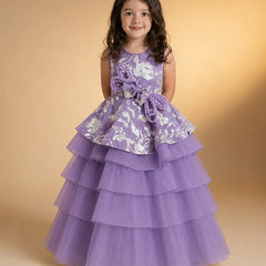 A young girl with dark curly hair wearing a lavender purple formal party dress with white floral embroidery and a tiered tulle skirt, standing against a warm beige studio background.