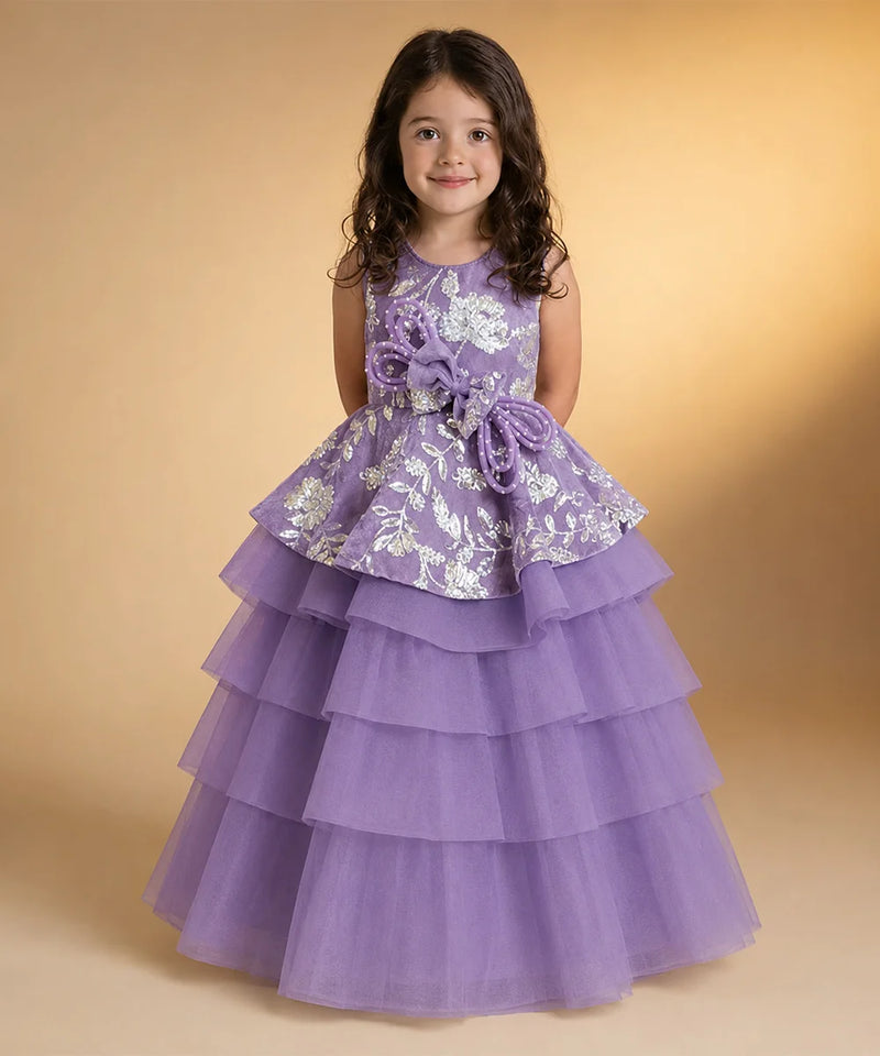 A young girl with dark curly hair wearing a lavender purple formal party dress with white floral embroidery and a tiered tulle skirt, standing against a warm beige studio background.