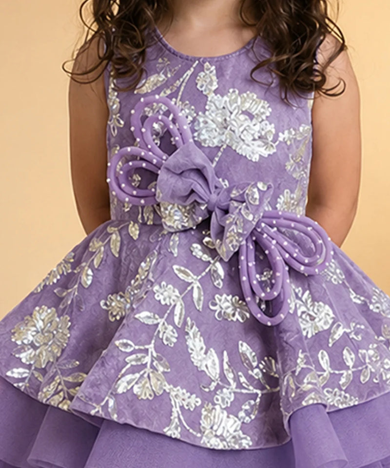 A sleeveless purple dress with a silver floral pattern, featuring a large purple bow at the waist, worn by a child against a solid beige background.