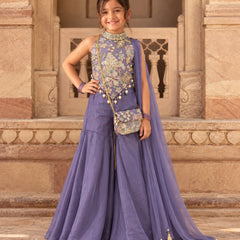 A young girl modeling a lavender purple traditional Indian outfit with a heavily embroidered floral bodice, matching skirt, and a sheer dupatta draped over her shoulder, standing in front of a stone architectural background.