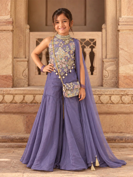 A young girl modeling a lavender purple traditional Indian outfit with a heavily embroidered floral bodice, matching skirt, and a sheer dupatta draped over her shoulder, standing in front of a stone architectural background.