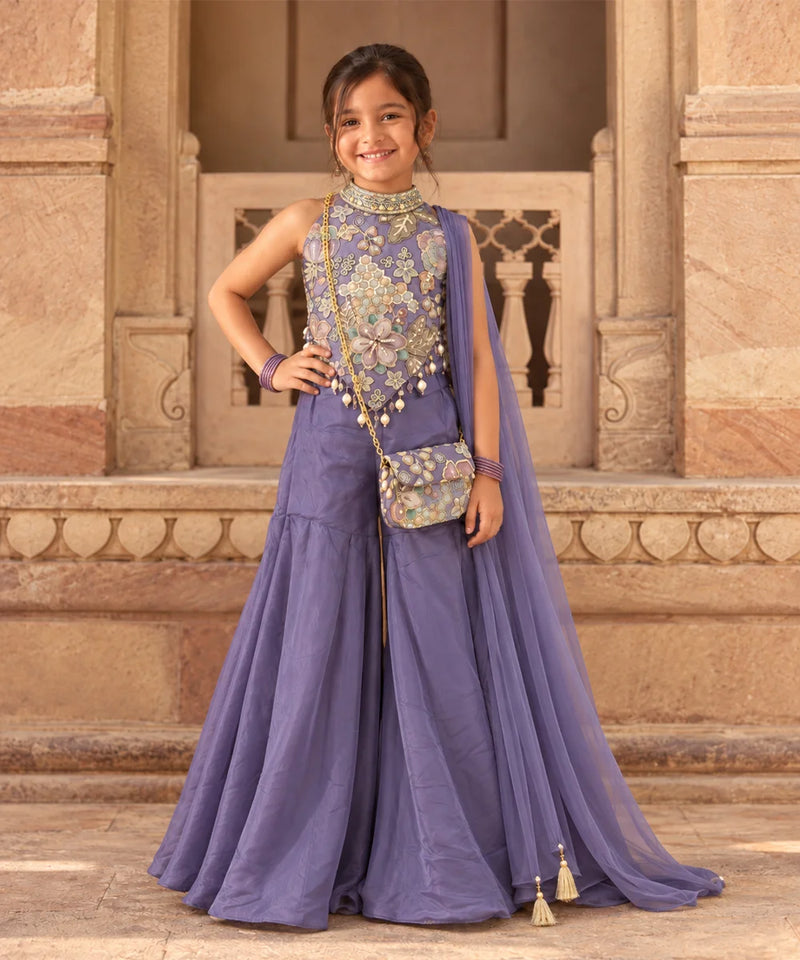 A young girl modeling a lavender purple traditional Indian outfit with a heavily embroidered floral bodice, matching skirt, and a sheer dupatta draped over her shoulder, standing in front of a stone architectural background.