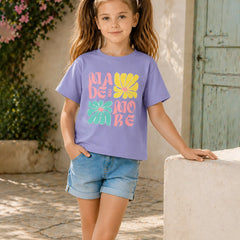 A young girl with pigtails stands outdoors wearing a lavender-purple graphic t-shirt with colorful retro text and flower designs, paired with denim shorts.