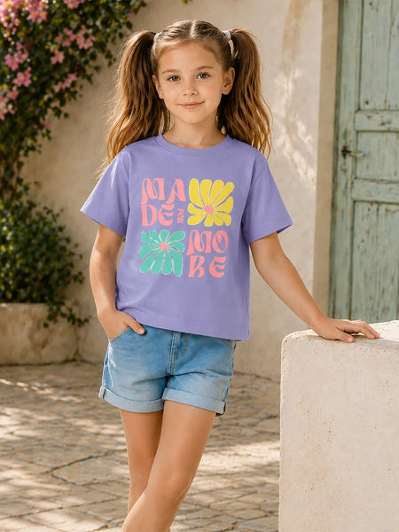 A young girl with pigtails stands outdoors wearing a lavender-purple graphic t-shirt with colorful retro text and flower designs, paired with denim shorts.