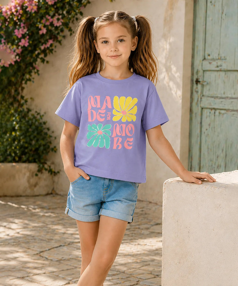 A young girl with pigtails stands outdoors wearing a lavender-purple graphic t-shirt with colorful retro text and flower designs, paired with denim shorts.