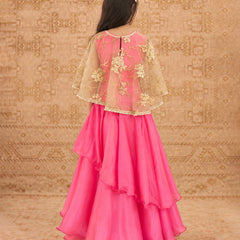 A woman viewed from behind wearing a bright pink traditional outfit with a layered flared skirt and a sheer beige embroidered cape-style overlay, posed against a patterned beige backdrop.