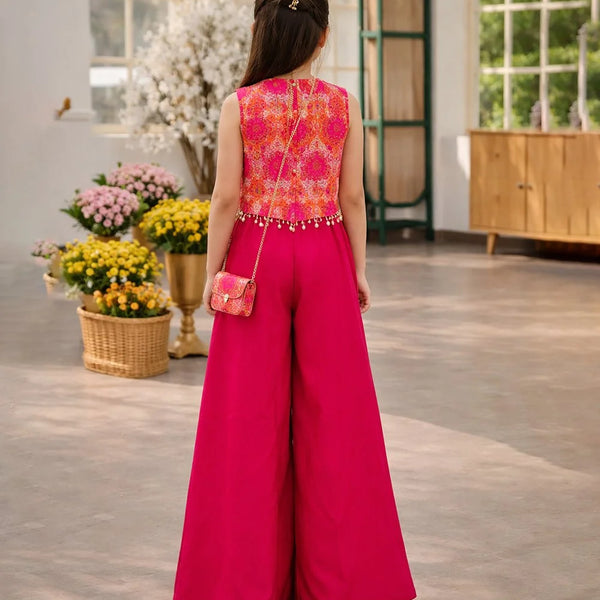 Woman in a bright pink outfit with a colorful top and wide-leg pants, standing in a room with flowers and a cabinet.