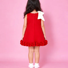 A young girl viewed from behind wearing a sleeveless red dress with a large white bow on the back shoulder and a ruffled hem, standing against a solid pink studio background.