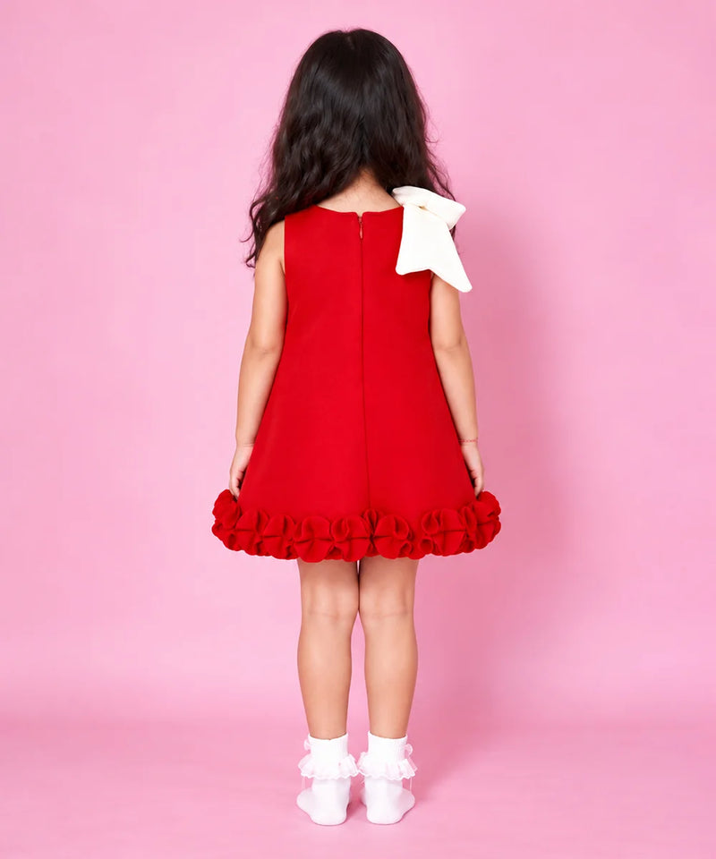 A young girl viewed from behind wearing a sleeveless red dress with a large white bow on the back shoulder and a ruffled hem, standing against a solid pink studio background.