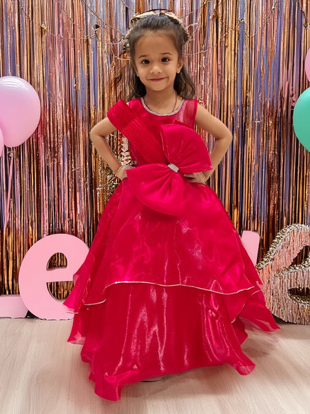 A young girl in a red party wear gown with a bow at the waist, standing in front of a decorative backdrop with balloons and the letters 'L O V E'.