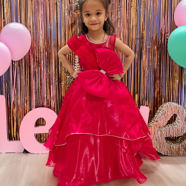 A young girl in a red party wear gown with a bow at the waist, standing in front of a decorative backdrop with balloons and the letters 'L O V E'.