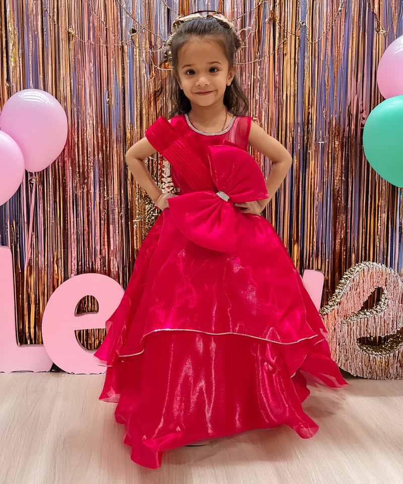 A young girl in a red party wear gown with a bow at the waist, standing in front of a decorative backdrop with balloons and the letters 'L O V E'.