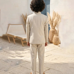 Person wearing a beige suit standing in a sunlit outdoor setting with decorative plants and a chair.