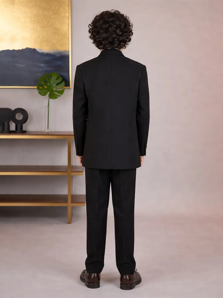 Young boy wearing a black traditional outfit with a decorative patch, standing against a neutral background.