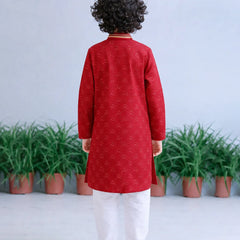 Child wearing a red kurta with white pants standing in front of green plants on a light gray background