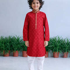 Child wearing a red kurta with white pants standing in front of green plants.