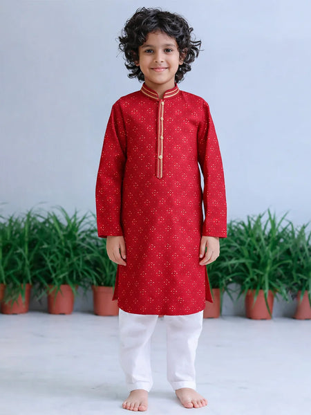Child wearing a red kurta with white pants standing in front of green plants.