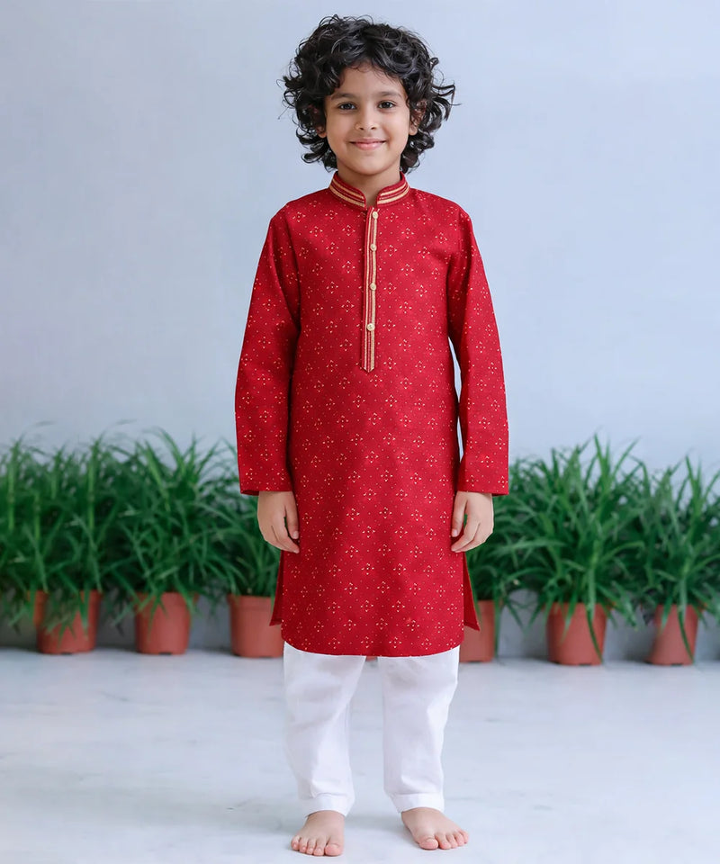 Child wearing a red kurta with white pants standing in front of green plants.