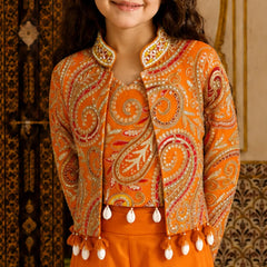 A young girl wearing a vibrant orange embroidered jacket with intricate paisley patterns and white tassel fringe, paired with matching orange trousers, set against a traditional patterned background.