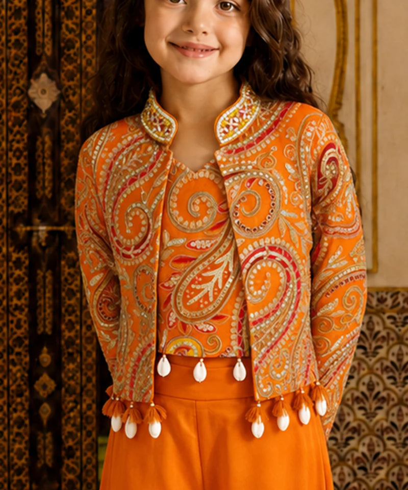 A young girl wearing a vibrant orange embroidered jacket with intricate paisley patterns and white tassel fringe, paired with matching orange trousers, set against a traditional patterned background.