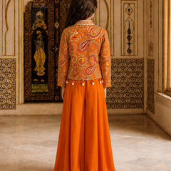 A woman viewed from behind wearing a traditional Indian outfit with an orange paisley embroidered jacket and a matching orange skirt, standing in an ornate interior room.