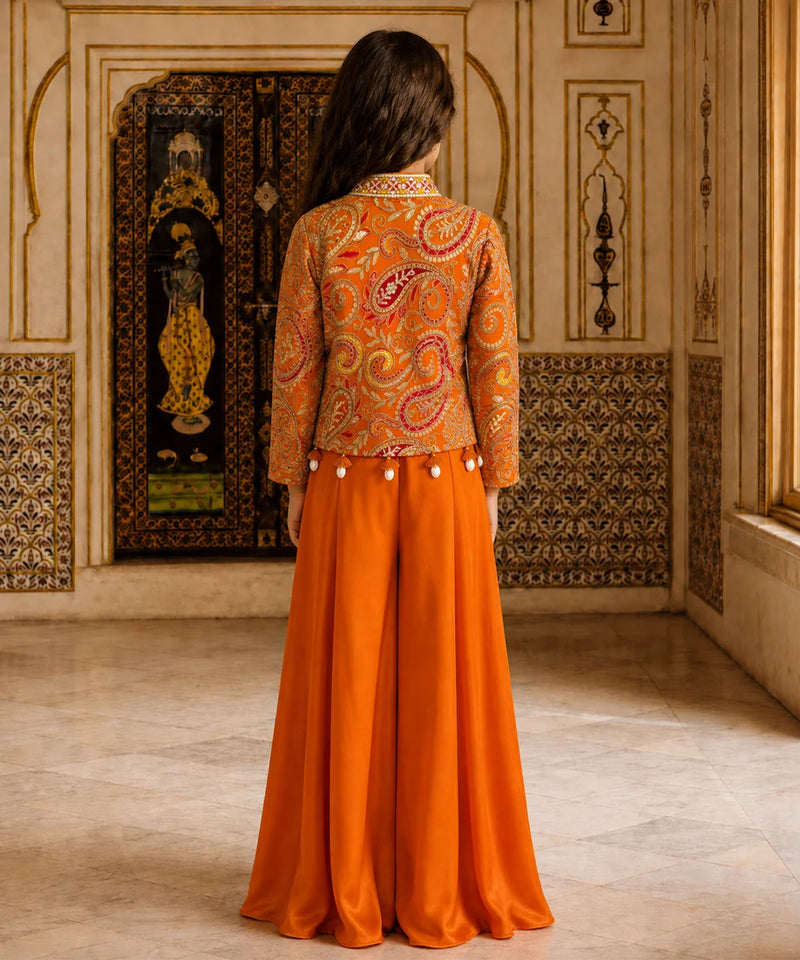 A woman viewed from behind wearing a traditional Indian outfit with an orange paisley embroidered jacket and a matching orange skirt, standing in an ornate interior room.