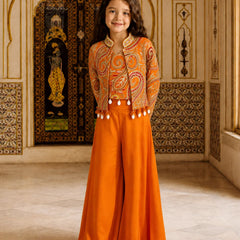 A young girl stands in an ornate indoor setting wearing a bright orange traditional outfit with a heavily embroidered cropped jacket and wide-leg pants. The background features decorative architectural details, patterned walls, and a large dark embroidered panel.