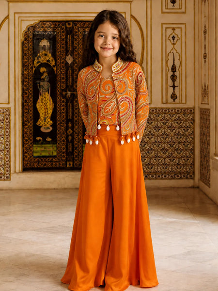 A young girl stands in an ornate indoor setting wearing a bright orange traditional outfit with a heavily embroidered cropped jacket and wide-leg pants. The background features decorative architectural details, patterned walls, and a large dark embroidered panel.