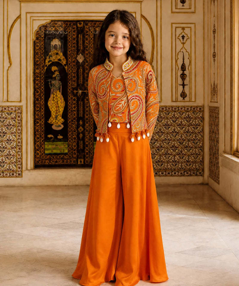A young girl stands in an ornate indoor setting wearing a bright orange traditional outfit with a heavily embroidered cropped jacket and wide-leg pants. The background features decorative architectural details, patterned walls, and a large dark embroidered panel.