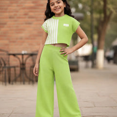 A young girl stands outdoors wearing a bright lime green two-piece outfit consisting of a short-sleeve crop top with white vertical stripes and matching wide-leg pants.