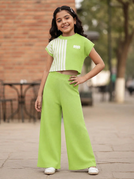 A young girl stands outdoors wearing a bright lime green two-piece outfit consisting of a short-sleeve crop top with white vertical stripes and matching wide-leg pants.