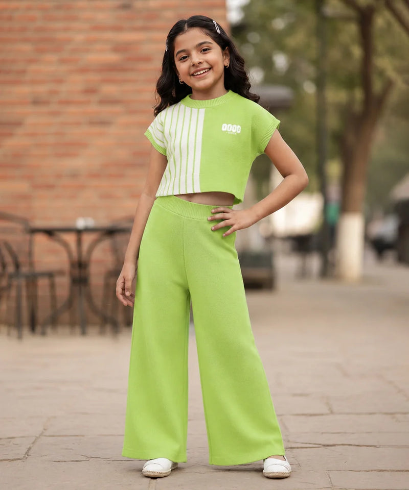 A young girl stands outdoors wearing a bright lime green two-piece outfit consisting of a short-sleeve crop top with white vertical stripes and matching wide-leg pants.