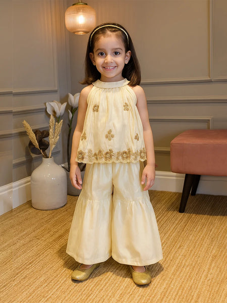Young girl in a cream dress with gold accents standing indoors.