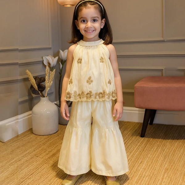 Young girl in a cream dress with gold accents standing indoors.