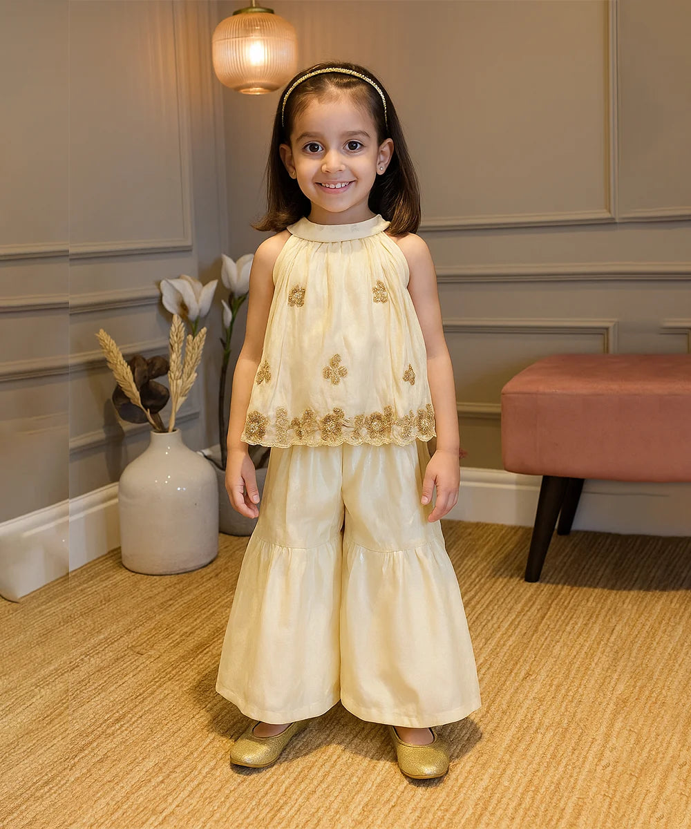 Young girl in a cream dress with gold accents standing indoors.