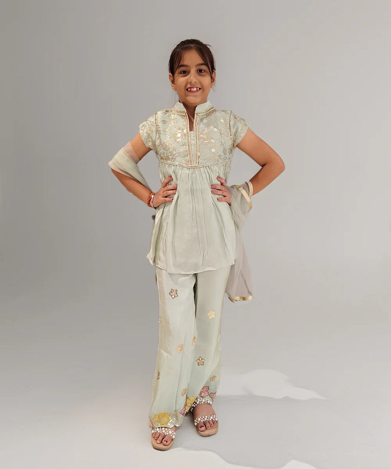 Young girl wearing a light green traditional outfit with floral patterns on a plain background