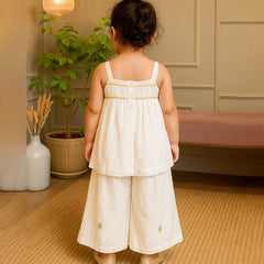 Child wearing a white dress with floral embroidery in a room with a plant and lamp.
