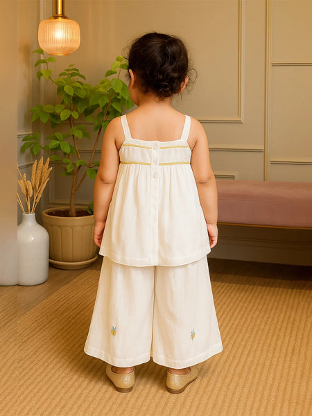 Child wearing a white dress with floral embroidery in a room with a plant and lamp.
