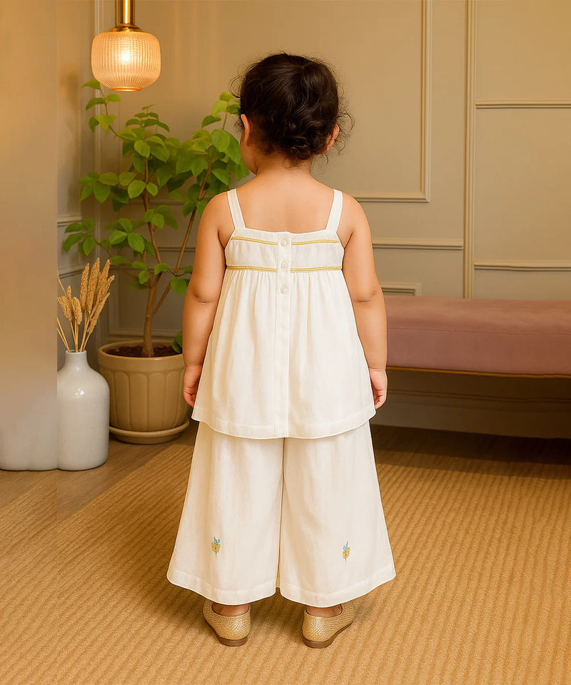 Child wearing a white dress with floral embroidery in a room with a plant and lamp.