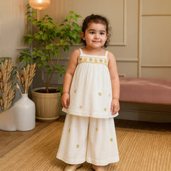 cute girl in a white dress with gold embroidery standing in a room with plants and furniture.