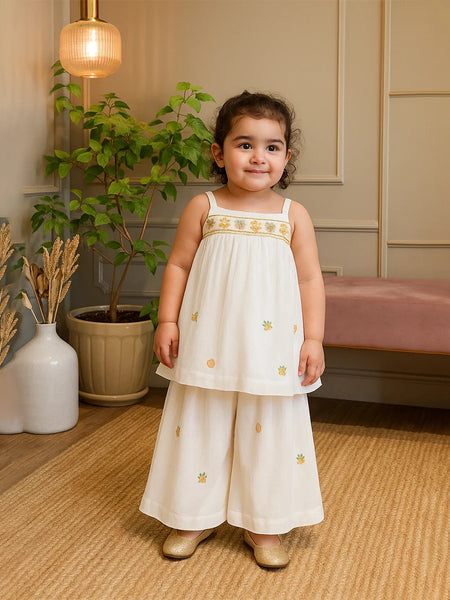 cute girl in a white dress with gold embroidery standing in a room with plants and furniture.